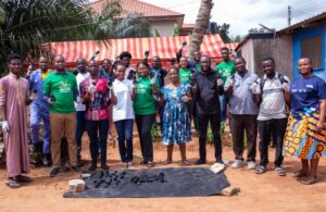 A group of young people, some wearing green WIN t-shirts, stand smiling and holding up small, black briquettes in their hands. A pile of the finished briquettes sits on the ground in front of them.